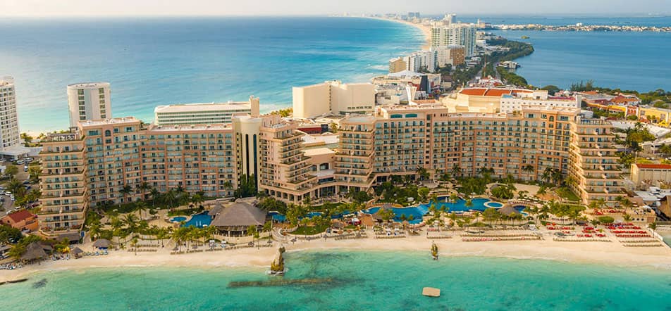 Aerial view of a large beachfront conference resort in Mexico, featuring multiple pools, palm trees, and sandy beach along a turquoise ocean, with cityscape and coastline extending into the distance—ideal for meetings and events.
