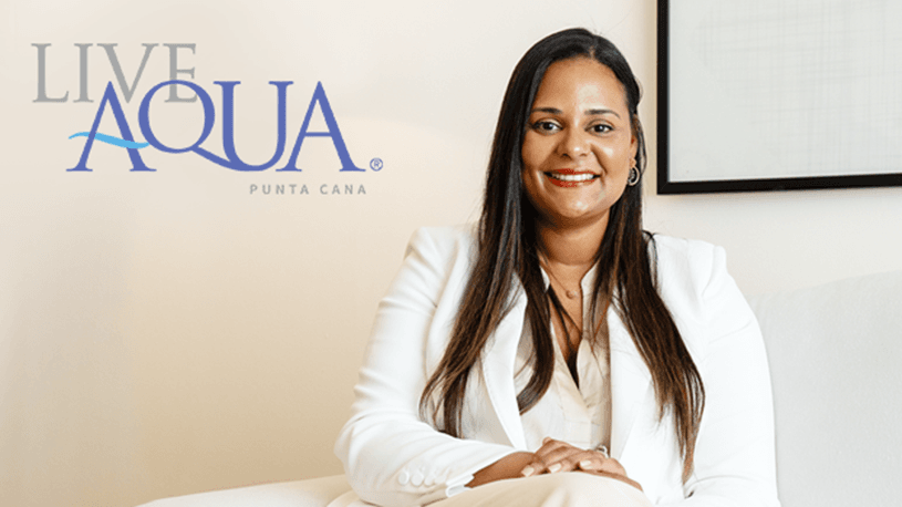 A woman in a white suit sits on a white sofa, smiling. The Live Aqua Punta Cana logo appears on the wall behind her—a bright, minimalistic setting that reflects the sophisticated ambiance found at Mexico’s top resorts and hotels for meetings and conference events.