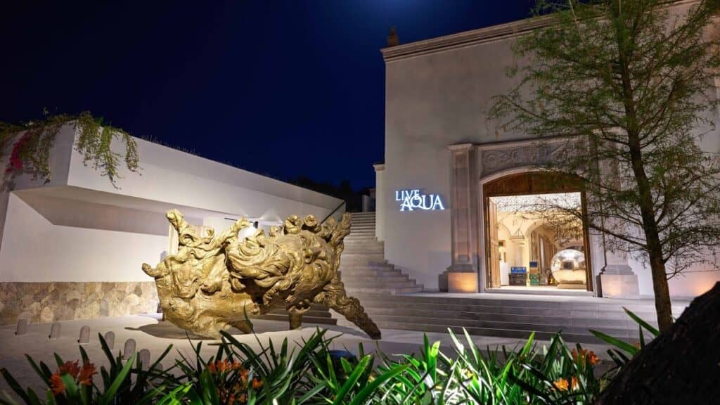 A large abstract bronze sculpture stands outside the well-lit Live Aqua building at night, surrounded by greenery and grand stairs—a striking entrance to one of Mexico’s premier resorts for meetings and conference events.
