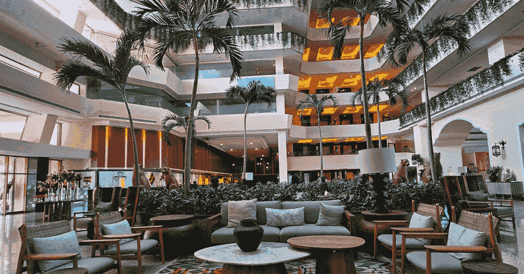 A spacious hotel lobby in Mexico, featuring tall palm trees, modern sofas and chairs around a coffee table—perfect for meetings or conference events. Multiple floors with balconies and greenery above, plus warm lighting, create a welcoming atmosphere.