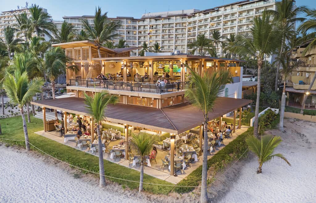 A two-story beachfront restaurant with outdoor seating surrounded by palm trees, diners enjoying meals, and a large hotel building in the background—perfect for meetings or conference events at Mexico resorts along a sandy beach.