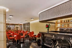 Moderno bar lounge con sillas rojas y pequeñas mesas redondas, suelos de mármol negro, barra de mármol negro, motivos geométricos en las paredes y estantes con bebidas: perfecto para reuniones o eventos en los mejores hoteles y resorts de México. Un jarrón con flores rojas adorna la barra.