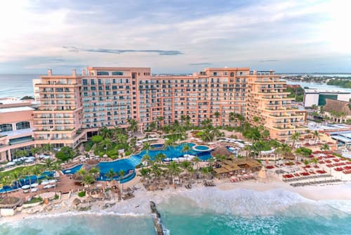 Aerial view of a large beachfront resort in Mexico with multiple pools, palm trees, lounge chairs, and umbrellas—perfect for meetings or conference events—set along a sandy shoreline with turquoise ocean waves under a partly cloudy sky.