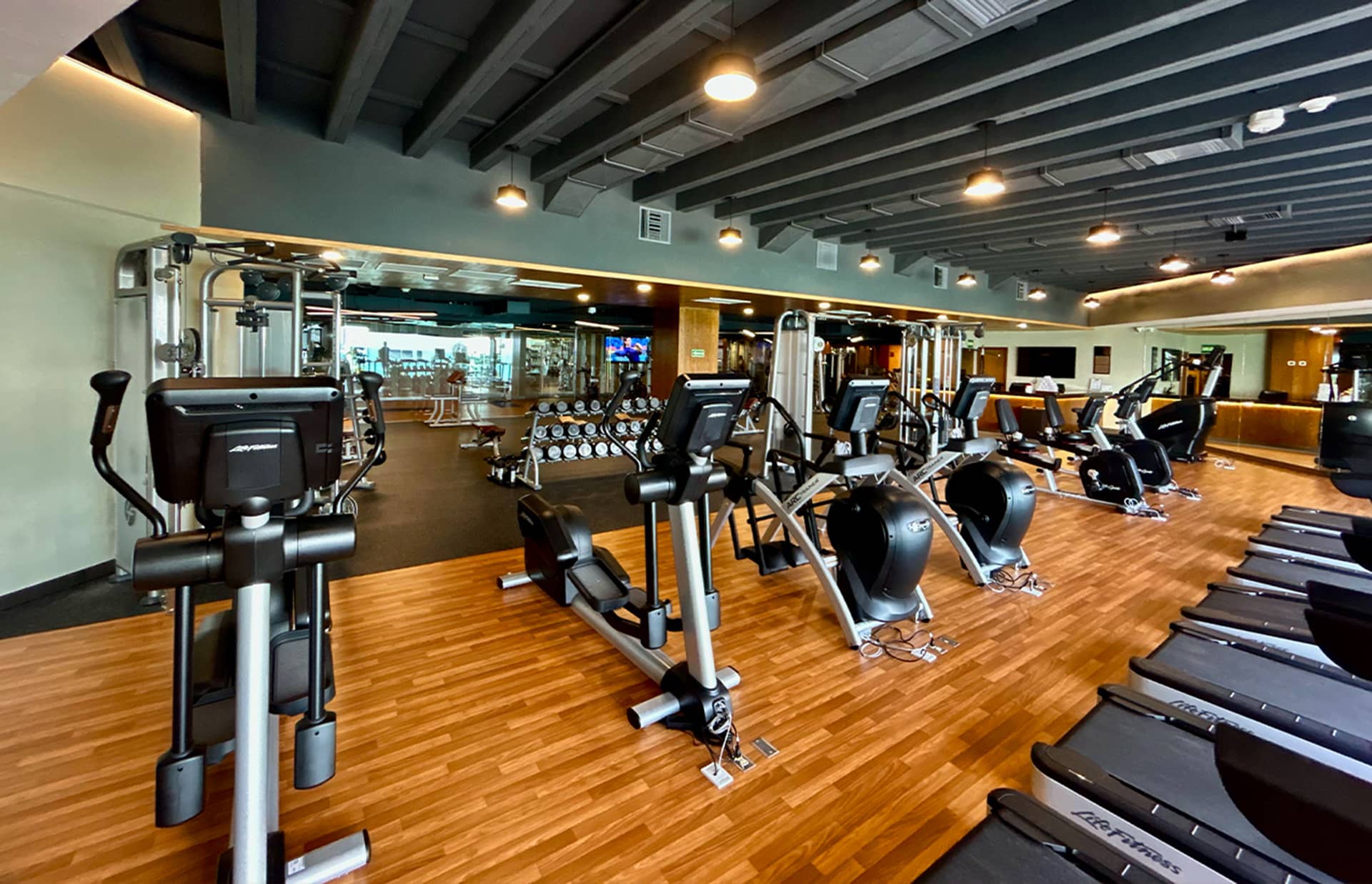 A modern gym with wood flooring, featuring treadmills, elliptical machines, weight machines, and free weights. Ideal for guests attending conference events at Mexico resorts or hotels, the spacious workout area is bright and exceptionally clean.