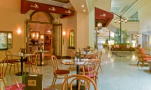 Una acogedora cafetería de hotel con mesas redondas y sillas de madera, situada cerca del vestíbulo, con iluminación brillante, plantas y cómodas zonas de estar; ideal para reuniones informales en hoteles de México o para relajarse después de eventos de conferencias.