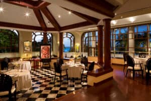 Elegante restaurante con suelos de baldosas blancas y negras, mesas redondas con manteles blancos, sillas de madera oscura y grandes ventanales con vistas a la puesta de sol: perfecto para eventos o reuniones en los mejores hoteles y complejos turísticos de México.