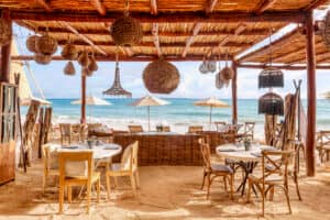 Restaurante a pie de playa con mesas y sillas de madera sobre la arena, a la sombra de un techo de paja. Perfecto para reuniones informales o eventos de conferencias, con vistas al océano, es ideal para los huéspedes que se alojan en los hoteles y complejos turísticos cercanos de México.