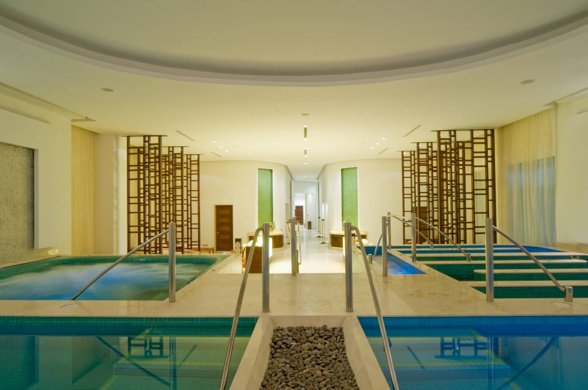 Modern indoor spa room with pools and hot tubs, metal handrails, wooden screens, and soft lighting. This serene space is perfect for relaxation and wellness during conference events at resorts or hotels in Mexico.
