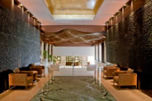 El vestíbulo de un hotel moderno, con sillones marrones alrededor de mesas de cristal, paredes oscuras con textura, un panel de suelo de cristal estampado y grandes ventanales, da la bienvenida a los huéspedes de uno de los complejos turísticos más elegantes de México, perfecto para reuniones o eventos de conferencias en un entorno espacioso y con luz natural.