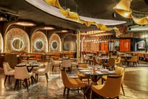 Un interior de restaurante moderno, ideal para reuniones, con iluminación cálida, sillas de color beige y naranja, mesas redondas preparadas para comer, paredes de ladrillo visto, grandes espejos redondos y singulares decoraciones de techo en forma de hojas doradas; perfecto para eventos de conferencias.