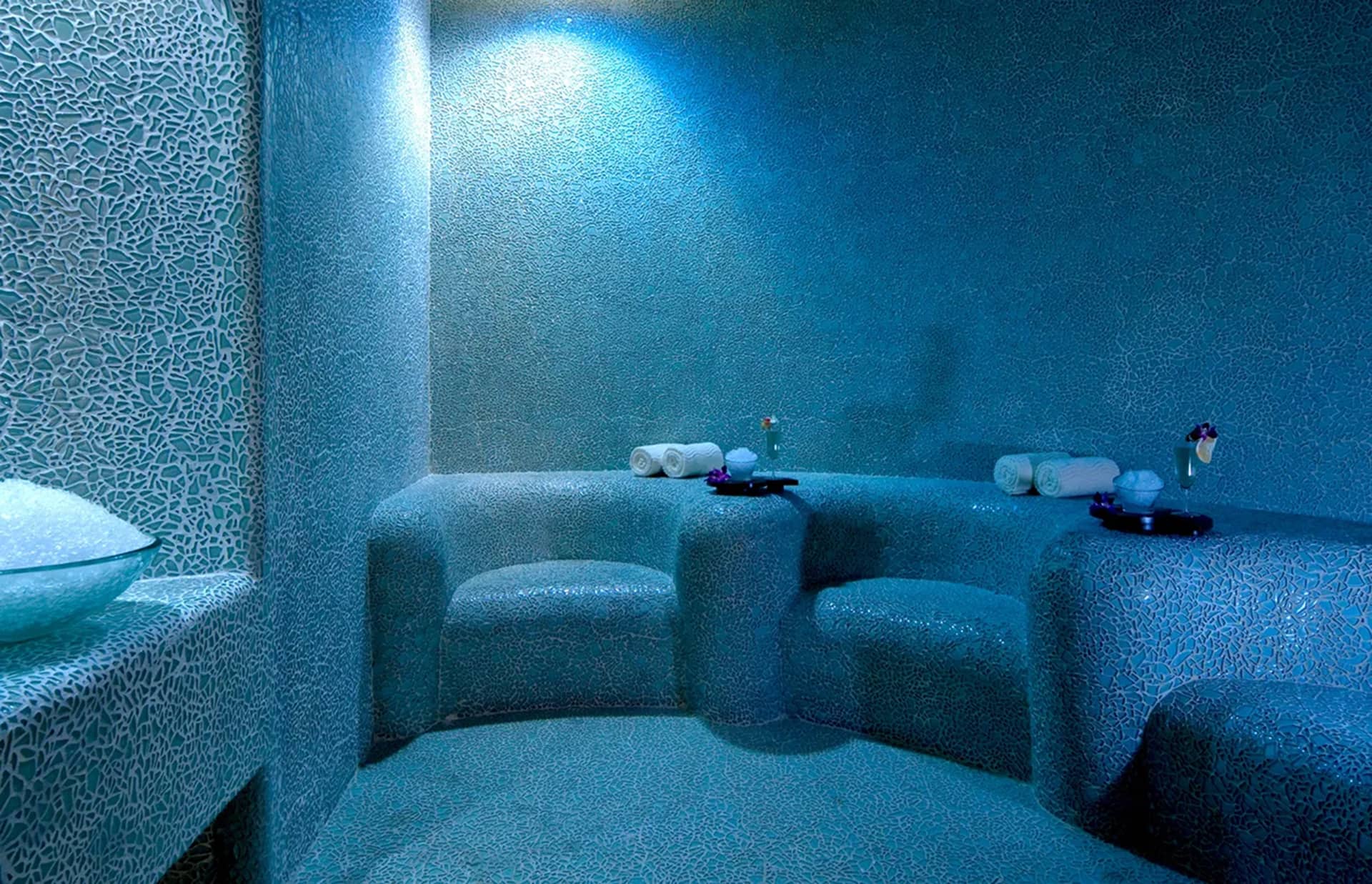 A modern steam room at one of Mexico's premier resorts, featuring curved blue mosaic-tiled benches, neatly rolled white towels, spa trays, and a bowl of stones—all under soft lighting—perfect for relaxing after meetings or conference events.