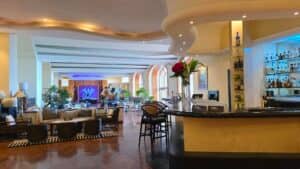 Un elegante salón y bar de hotel en uno de los mejores complejos turísticos de México, con una decoración moderna, una barra curva, estantes con botellas de licor, amplias zonas de estar para reuniones o eventos de conferencias, grandes ventanales arqueados y exuberantes plantas por todas partes.