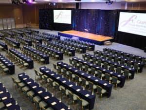 El amplio salón de conferencias del Grand Fiesta Americana Coral Beach Cancún, ideal para reuniones y eventos, está acondicionado con filas de mesas y sillas frente a un escenario con dos pantallas de proyección. En cada mesa hay blocs de notas y botellas de agua.