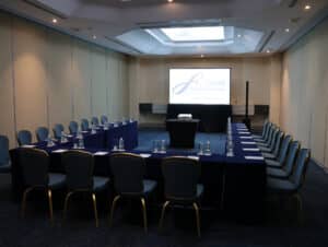 La sala de conferencias del Grand Fiesta Americana Coral Beach Cancun Resort & Spa cuenta con una mesa en forma de U, sillas azules, botellas de agua, blocs de notas y una pantalla de proyección. Este espacio hotelero en México está bien iluminado gracias a una claraboya, ideal para eventos y conferencias.