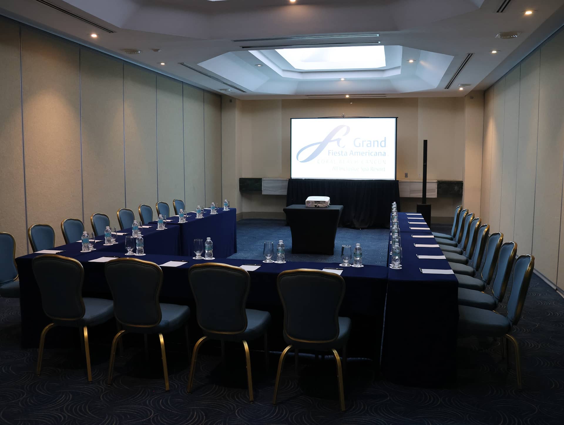 A conference room for meetings at Grand Fiesta Americana Coral Beach Cancun Resort & Spa features a U-shaped table, blue chairs, water bottles, notepads, and a projector screen. This Mexico hotel venue is well-lit with a skylight, ideal for conference events.