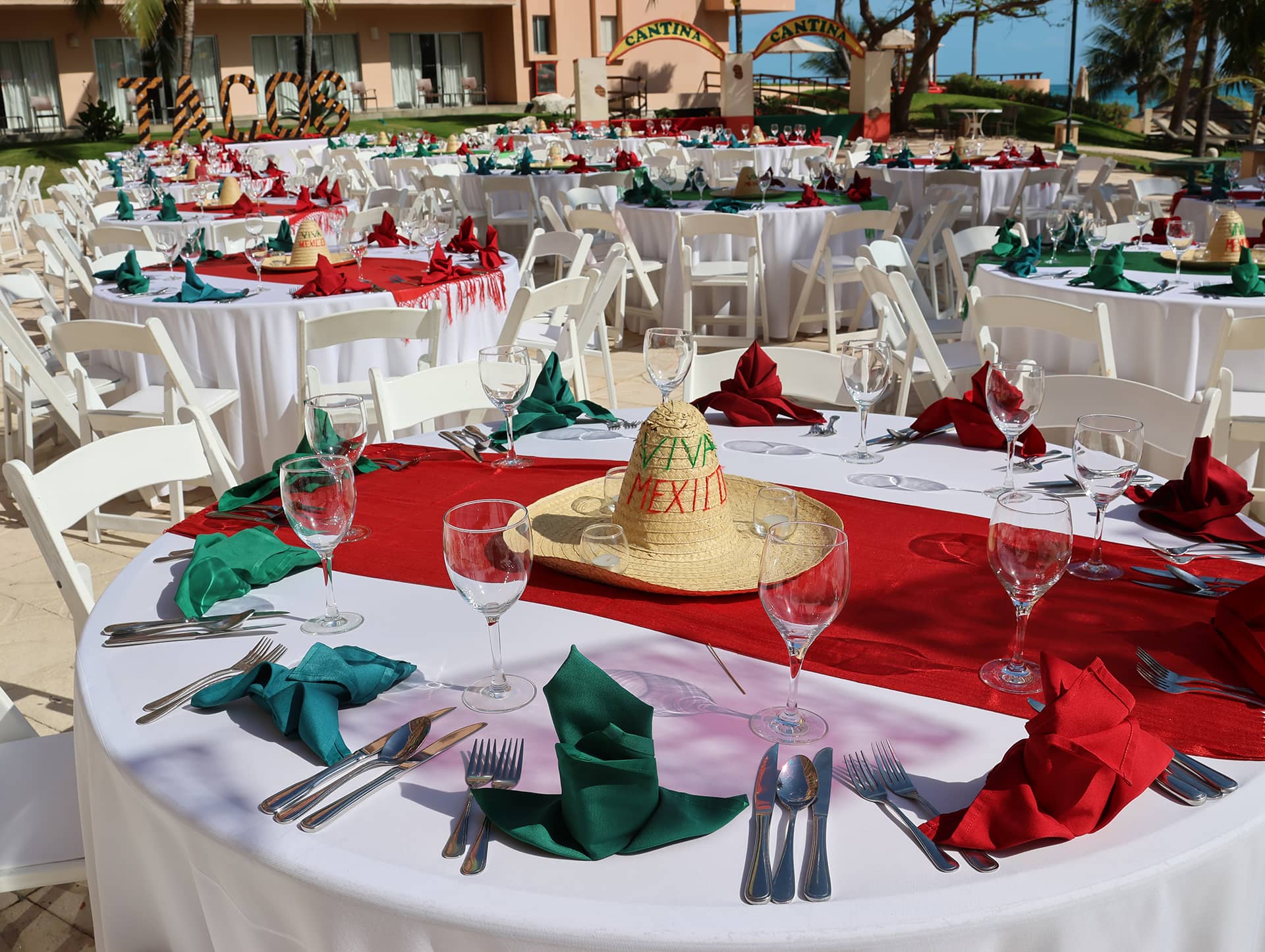 Round tables set with white tablecloths, red and green napkins, and glassware for conference events at Mexico resorts; a straw sombrero reading Viva Mexico decorates one table, with a building and palm trees in the background.
