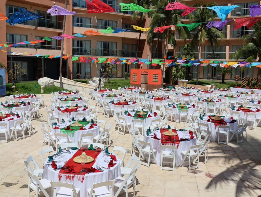Colorful papel picado banners hang above round tables covered with white tablecloths and festive red and green decorations, set for an outdoor conference or meeting in a courtyard at a Mexico resort, with palm trees and a building in the background.