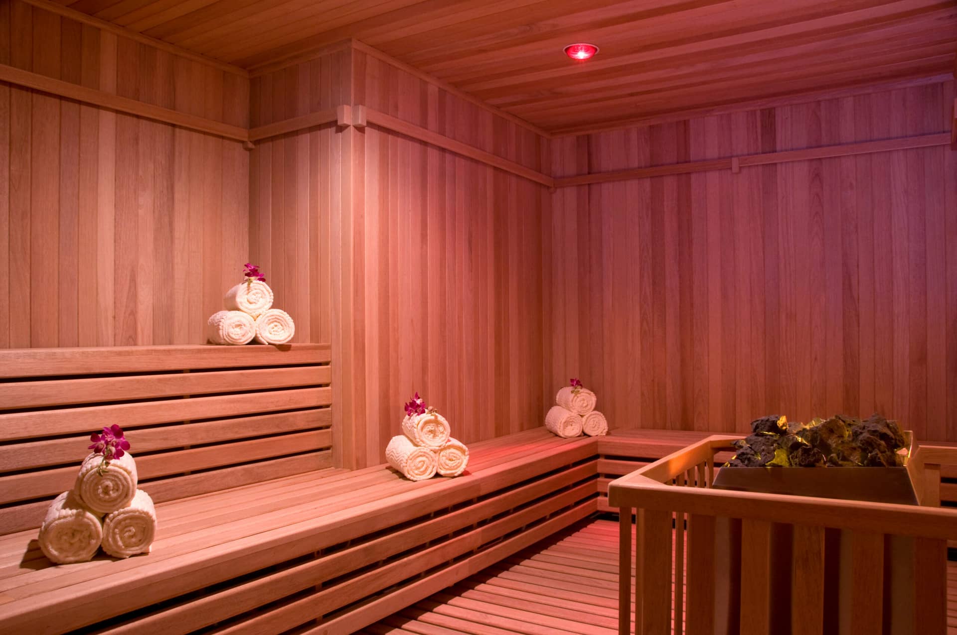 A wooden sauna room with benches, stacks of rolled white towels topped with purple flowers, and a basket of sauna leaves—all under warm, soft lighting—offers a tranquil retreat at luxury Mexico hotels and conference events resorts.