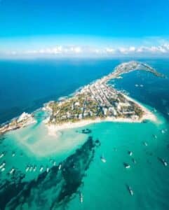 Vista aérea de una estrecha isla en México, rodeada de aguas turquesas, playas de arena y embarcaciones. La isla está bordeada de complejos turísticos y hoteles, con una exuberante vegetación y el profundo azul del océano que se extiende hasta el horizonte bajo un cielo despejado.