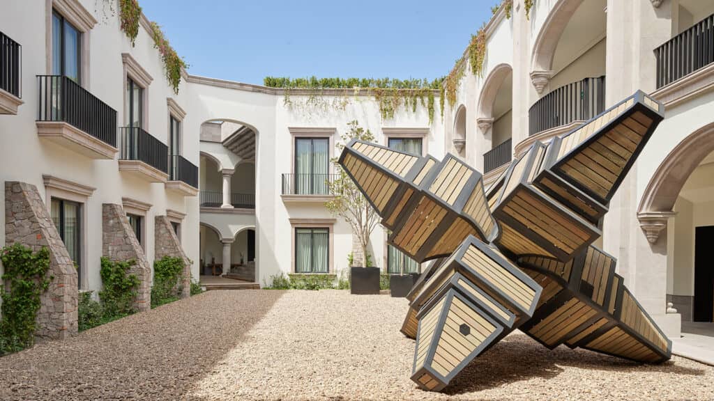 A modern courtyard at Mexico resorts with arched walkways, white walls, and balconies, featuring a large geometric wooden and metal sculpture on gravel. Greenery hangs from the roof and climbs the walls—perfect for meetings or conference events.