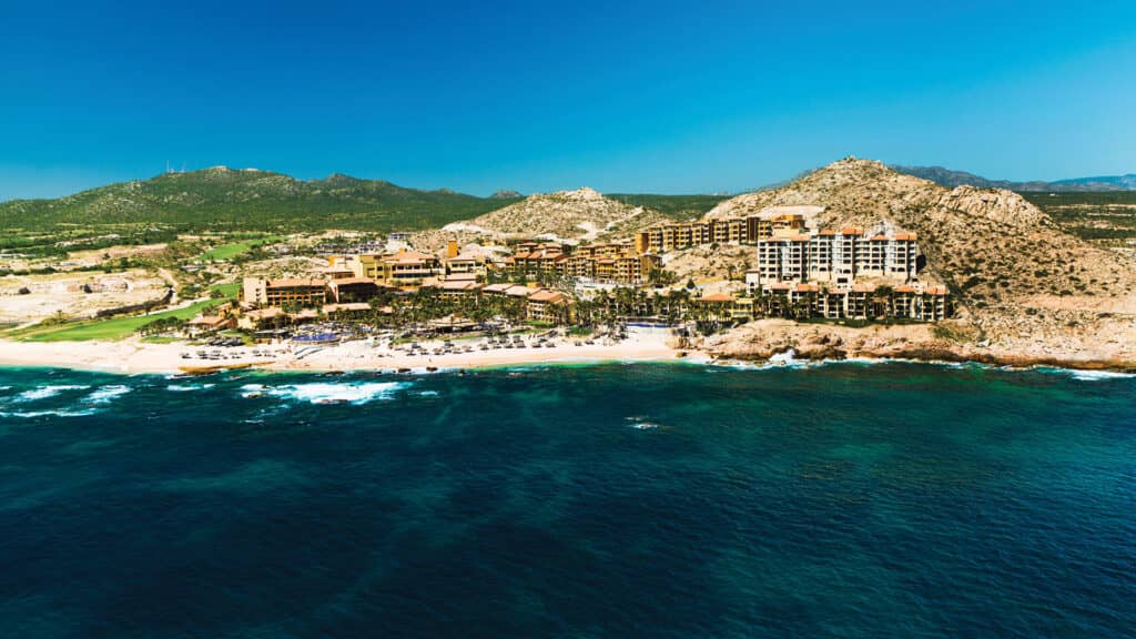 Aerial view of a beachfront resort in Mexico with multiple buildings nestled between sandy shores and rocky hills, perfect for conferences and meetings, bordered by turquoise ocean waves and surrounded by green landscape.