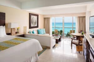 Habitación de hotel luminosa con una cama grande, zona de estar, mesa y TV, con ventanales del suelo al techo y balcón con vistas al mar; perfecta para relajarse después de reuniones o eventos en uno de los mejores resorts de México. Decoración moderna con sutiles toques tropicales.