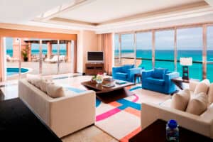 Un amplio y moderno salón con sofás color crema, sillones azules, una colorida alfombra y grandes ventanales con vistas a un patio con piscina y al océano; perfecto para relajarse después de reuniones en los mejores resorts de México. La luz natural inunda la estancia.