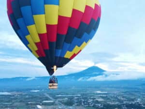 Un colorido globo aerostático con una cesta llena de gente flota sobre un paisaje verde cerca de hoteles en México, con un volcán humeante y un cielo nublado al fondo.