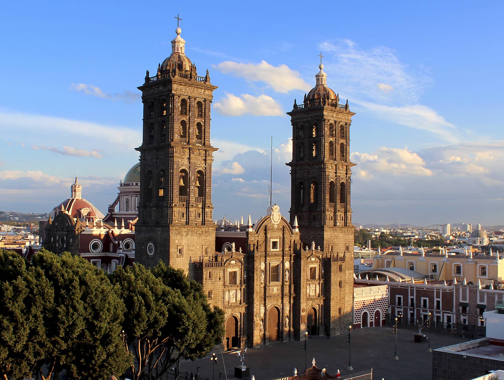 A view of Puebla Cathedral in Mexico, featuring two tall bell towers and ornate stone architecture, set against a blue sky with scattered clouds and surrounded by city buildings, trees, and nearby hotels perfect for meetings or conference events.