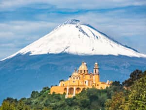 En primer plano, una iglesia amarilla con cúpulas se alza sobre una colina, rodeada de exuberante vegetación, con el volcán Popocatépetl, cubierto de nieve, elevándose majestuosamente al fondo: una vista inolvidable cerca de los mejores hoteles y centros de convenciones de México.