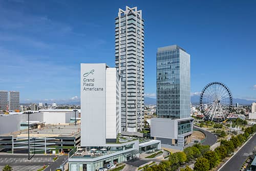 A modern cityscape featuring tall buildings, including the Grand Fiesta Americana hotel—one of Mexico’s top hotels for meetings and conference events—a Ferris wheel, and a multi-level parking structure under a clear blue sky.
