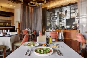 Un restaurante moderno y elegante en uno de los mejores resorts de México, con mesas preparadas para comer (ensalada, pan, vino, aceite y vinagre en primer plano); perfecto para reuniones o eventos de conferencias en medio de una decoración elegante y una iluminación tenue.