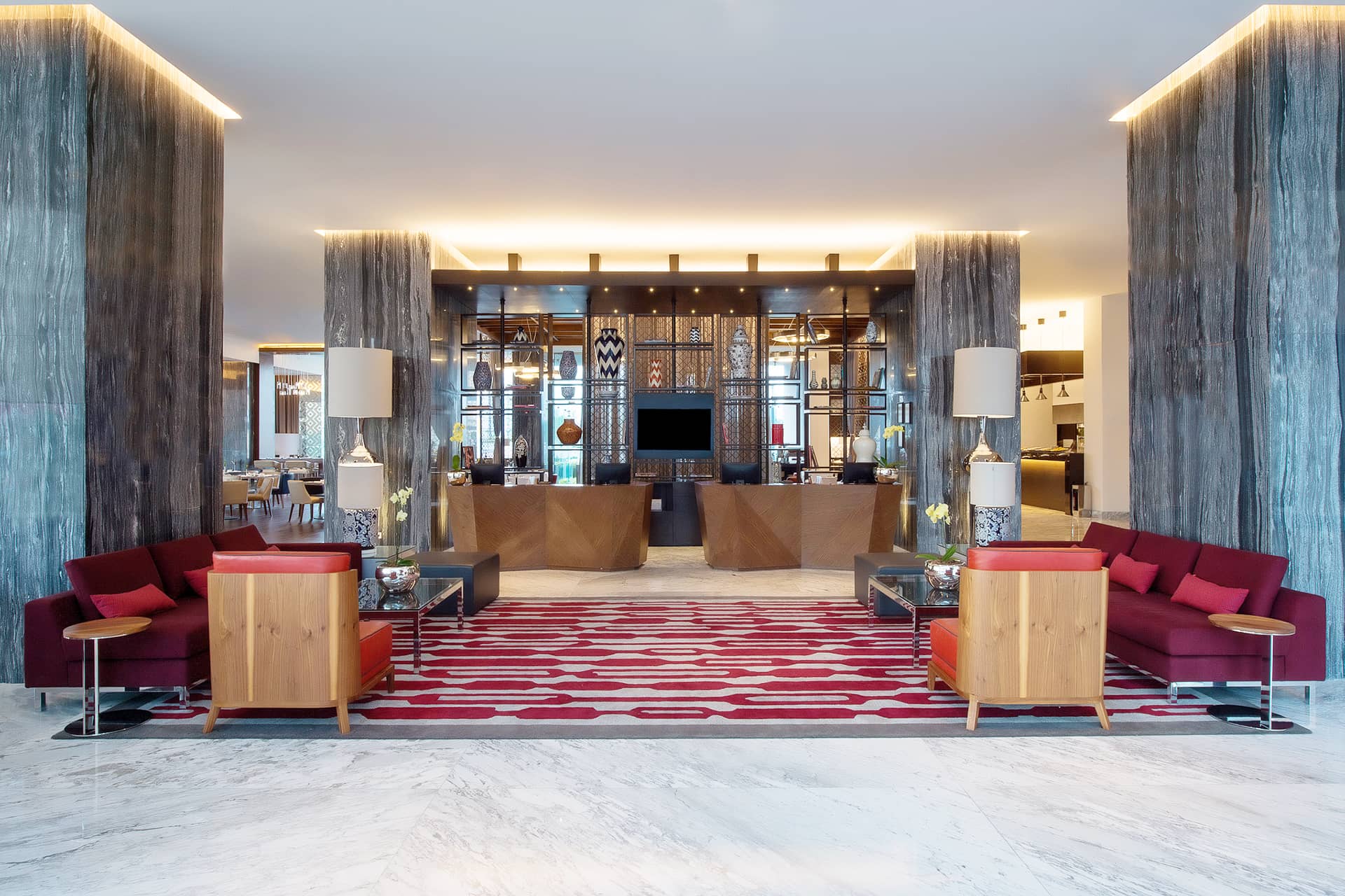 Modern hotel lobby with marble floors, red and wood sofas, geometric red pattern rug, and a reception desk in the center framed by two tall columns. Bright, spacious, and stylish interior design—ideal for meetings or conference events at Mexico hotels.