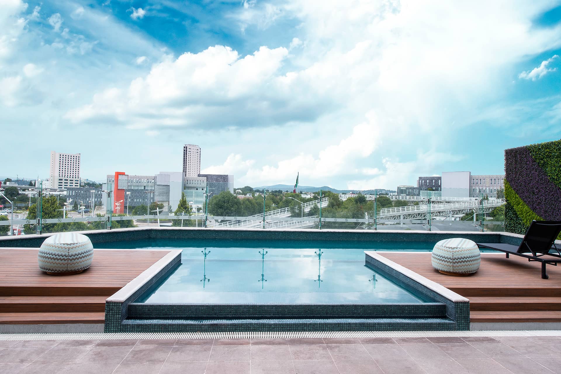A rooftop pool with clear water, wooden deck, two round poufs, and a lounge chair overlooks a cityscape with modern buildings—an ideal setting for conference events or meetings at Mexico hotels and resorts under a partly cloudy blue sky.