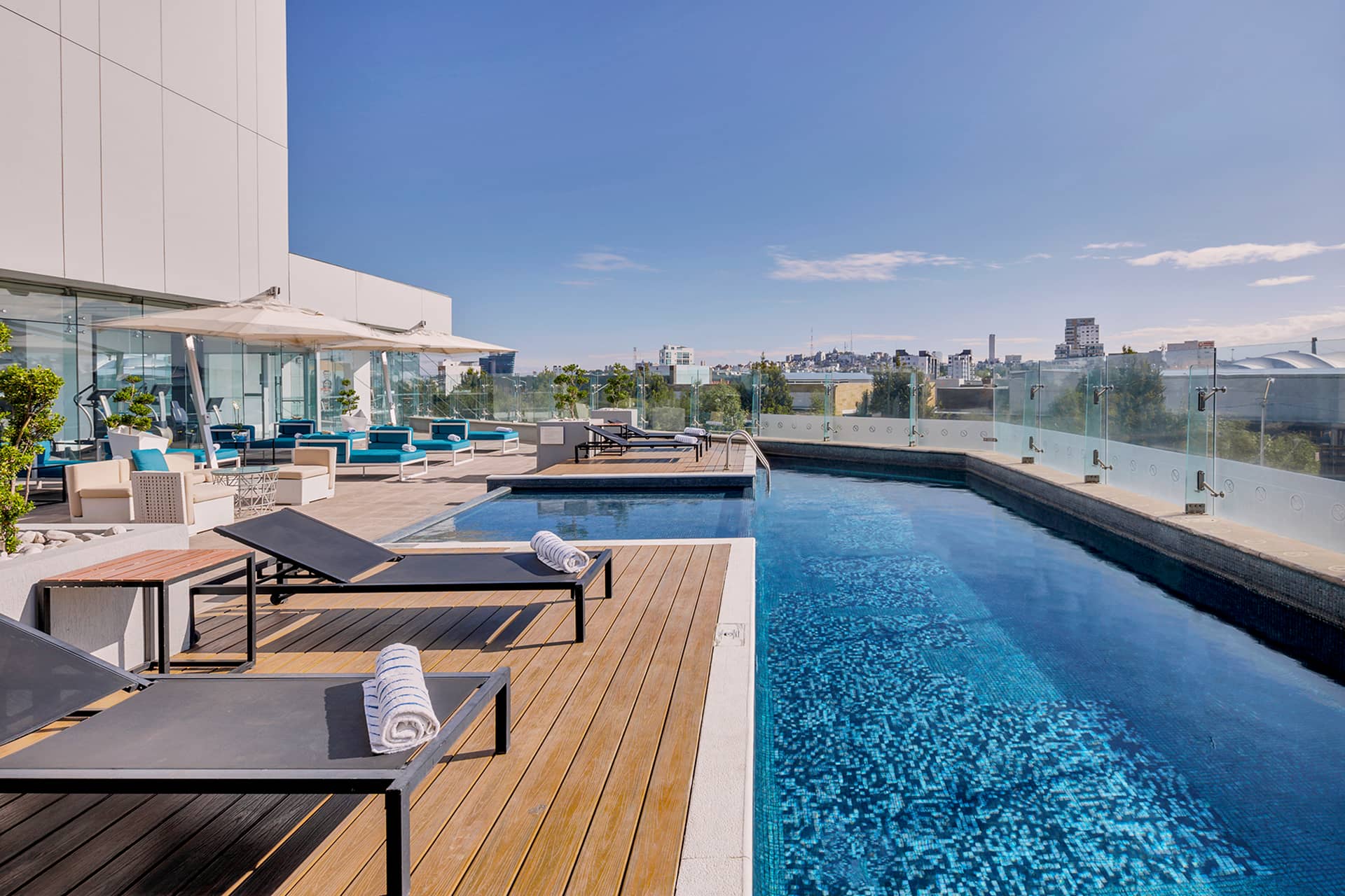 Rooftop swimming pool with lounge chairs and rolled towels, wooden deck, glass railing, and city skyline in the background—perfect for relaxing after conference events at top Mexico hotels and resorts under a clear blue sky.