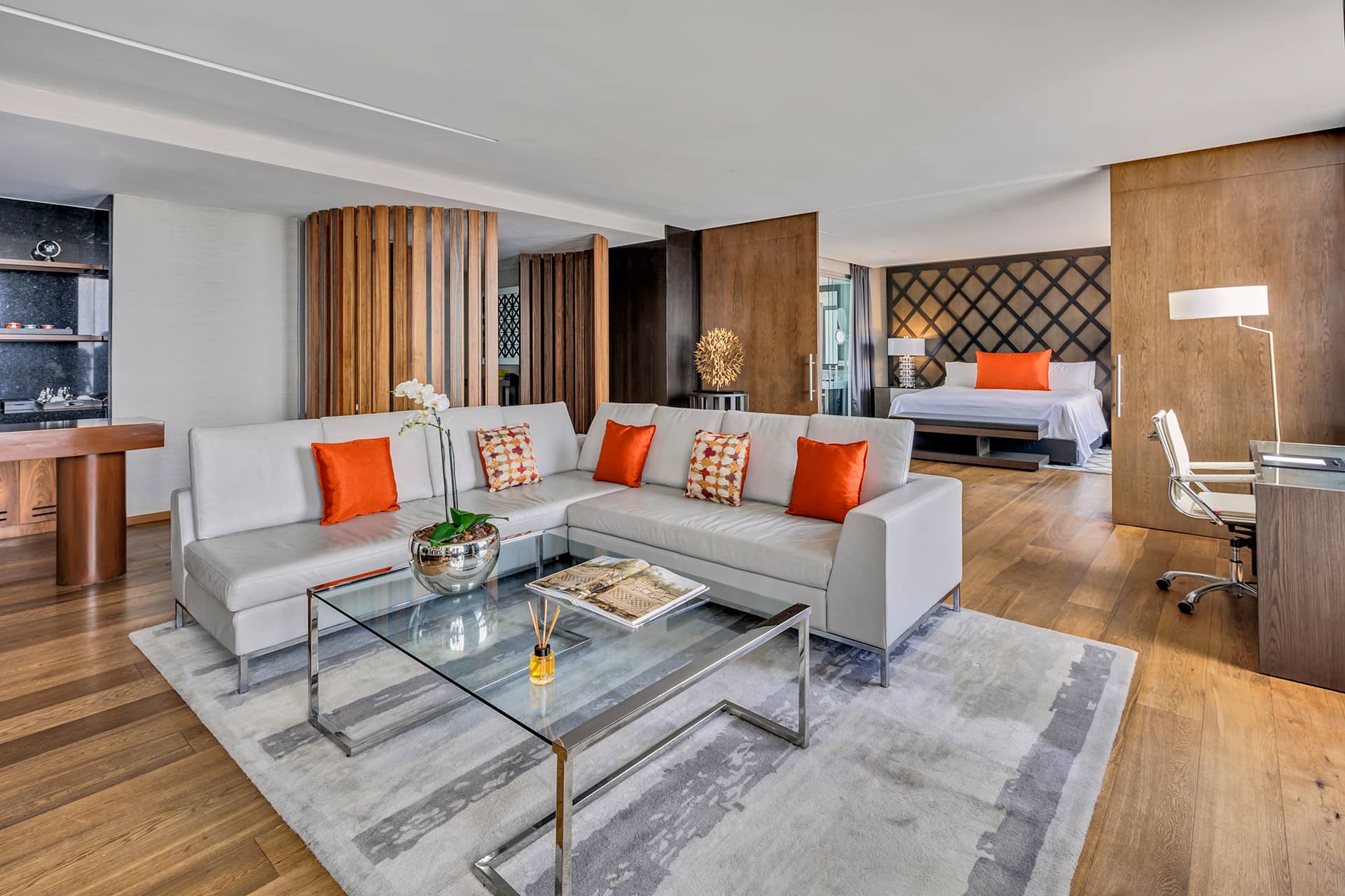 Modern open-plan suite ideal for meetings, featuring a gray sofa, orange accent pillows, glass coffee table, work desk, and visible bed. Wooden floors and walls, neutral tones, and natural light complete the stylish Mexico hotels decor.