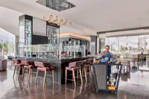 Un moderno bar de hotel con taburetes altos alrededor de una barra negra, iluminación elegante y grandes ventanales: perfecto para relajarse después de eventos o reuniones en los mejores hoteles de México. Un camarero uniformado prepara bebidas en un carrito móvil cerca del centro.