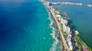 Vista aérea de una extensa costa con olas turquesas, playas de arena y altos edificios blancos que bordean la orilla. Al otro lado, se divisa una laguna con más hoteles y vegetación: un escenario ideal para eventos y conferencias en México.