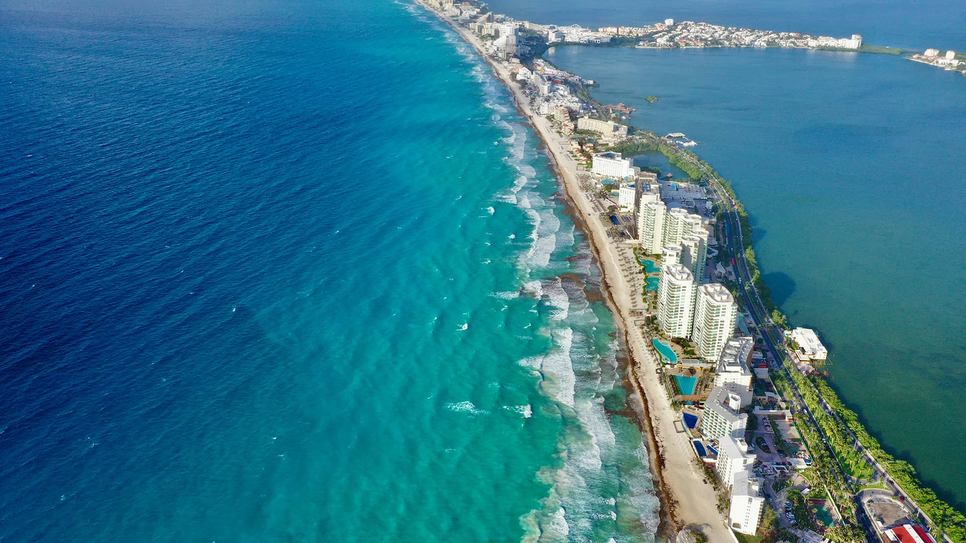 Aerial view of a long coastline with turquoise ocean waves, sandy beaches, and tall white buildings lining the shore. On the other side, a lagoon is visible with more hotels and greenery—an ideal setting for conference events in Mexico.