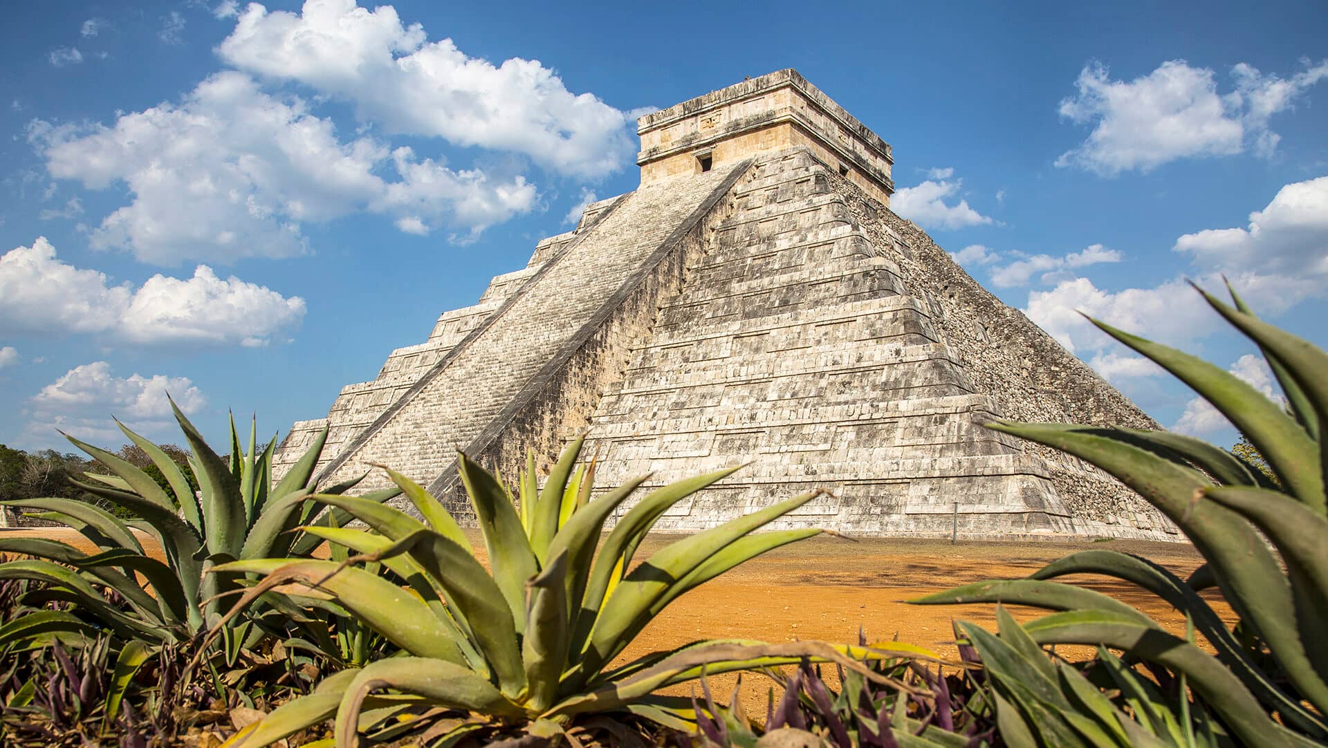 A large stone pyramid with steep steps, known as El Castillo, stands under a blue sky with scattered clouds. Succulent plants frame the ancient Mayan structure at Chichen Itza near some of Mexico’s top resorts and conference events destinations.