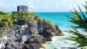 Una pequeña ruina de piedra antigua se alza sobre acantilados rocosos con vistas a las olas turquesas del océano y a una playa de arena, rodeada de exuberante vegetación bajo un cielo brillante y parcialmente nublado: una vista inspiradora perfecta para eventos y conferencias en los complejos turísticos de México.