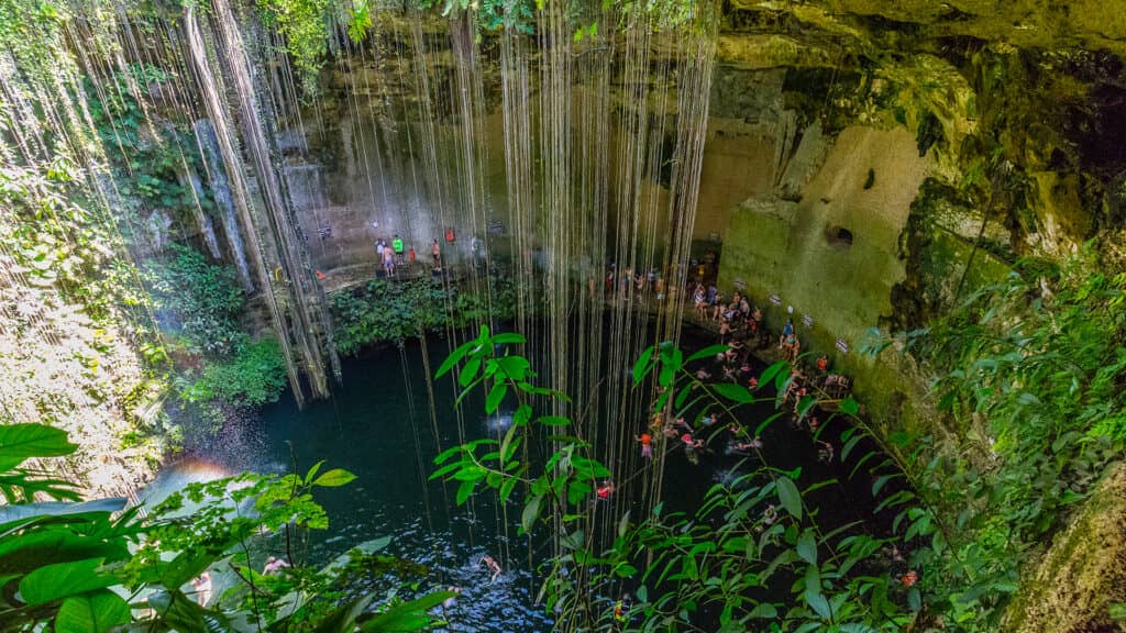 Lush green vines hang over the edge of a large, open cenote with clear water below. People gather and swim in the pool, surrounded by rocky cave walls and dense vegetation—an unforgettable escape at top Mexico resorts.