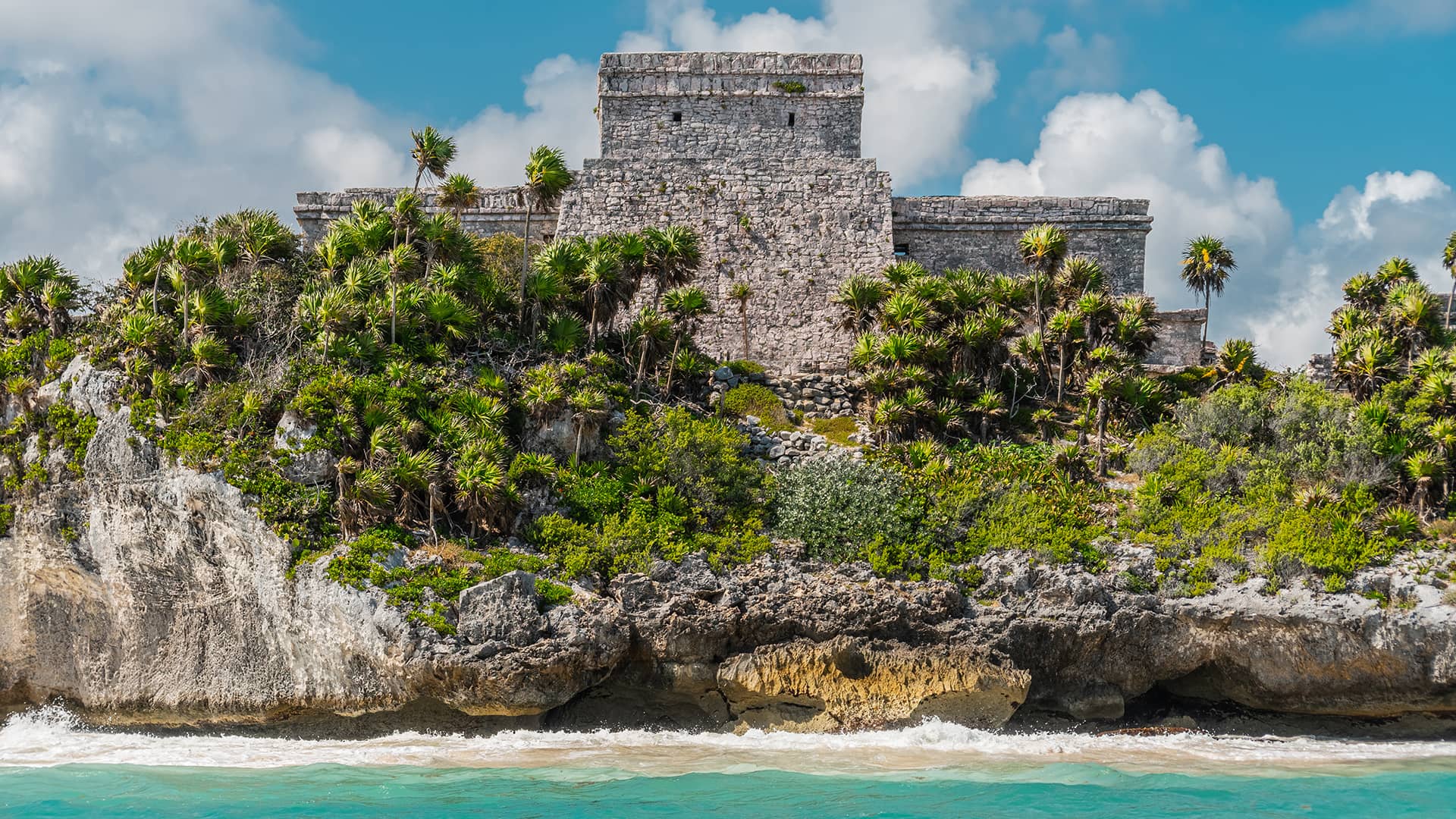 Ancient stone Mayan ruin atop a rocky cliff, surrounded by palm trees and greenery, overlooks turquoise ocean waters under a partly cloudy sky—a breathtaking view near top Mexico resorts and hotels perfect for meetings or conference events.