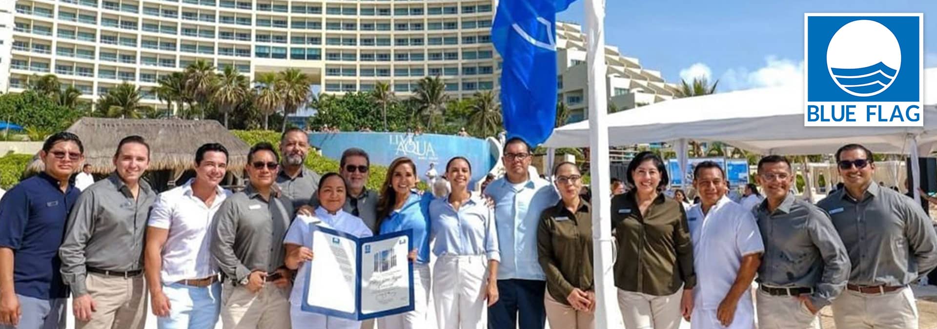 A group of people stand smiling outdoors near a beachside resort in Mexico, holding a certificate next to a raised blue flag. Palm trees and a large hotel building are in the background. A Blue Flag logo appears in the upper right corner.
