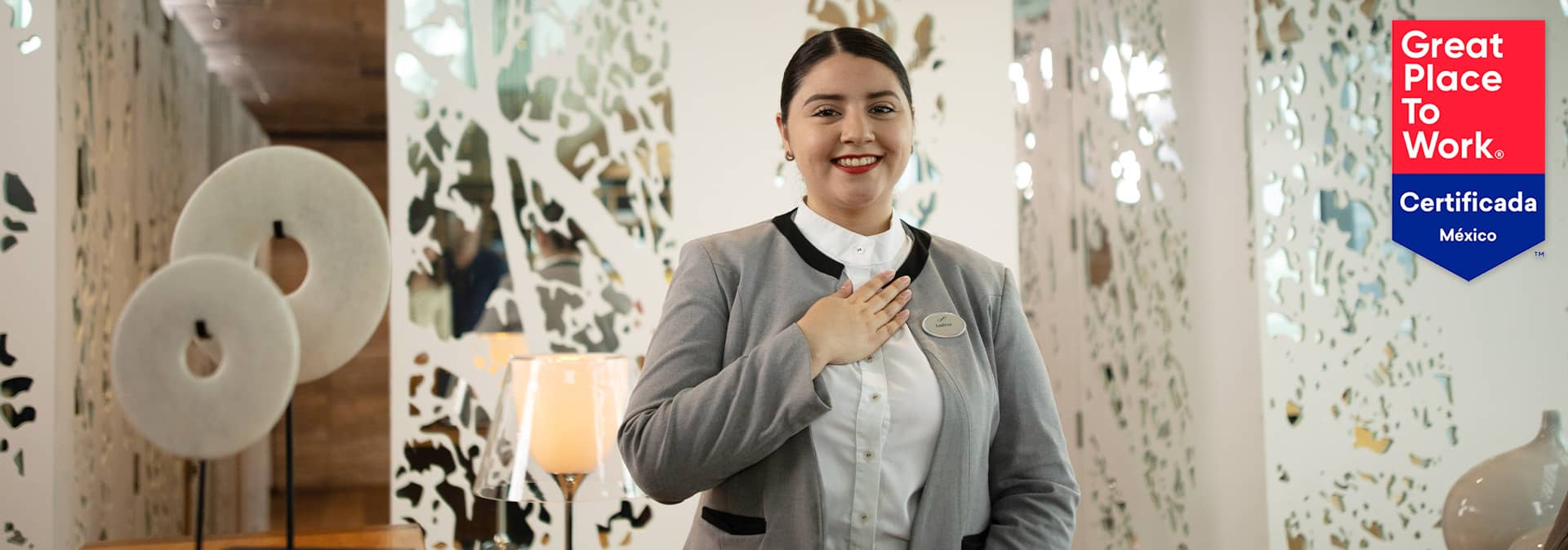 A woman in a gray blazer stands smiling with her hand on her chest in a modern office. On the right is a red and blue Great Place to Work Certificada México badge—a proud moment for Mexico hotels and conference events planners alike.