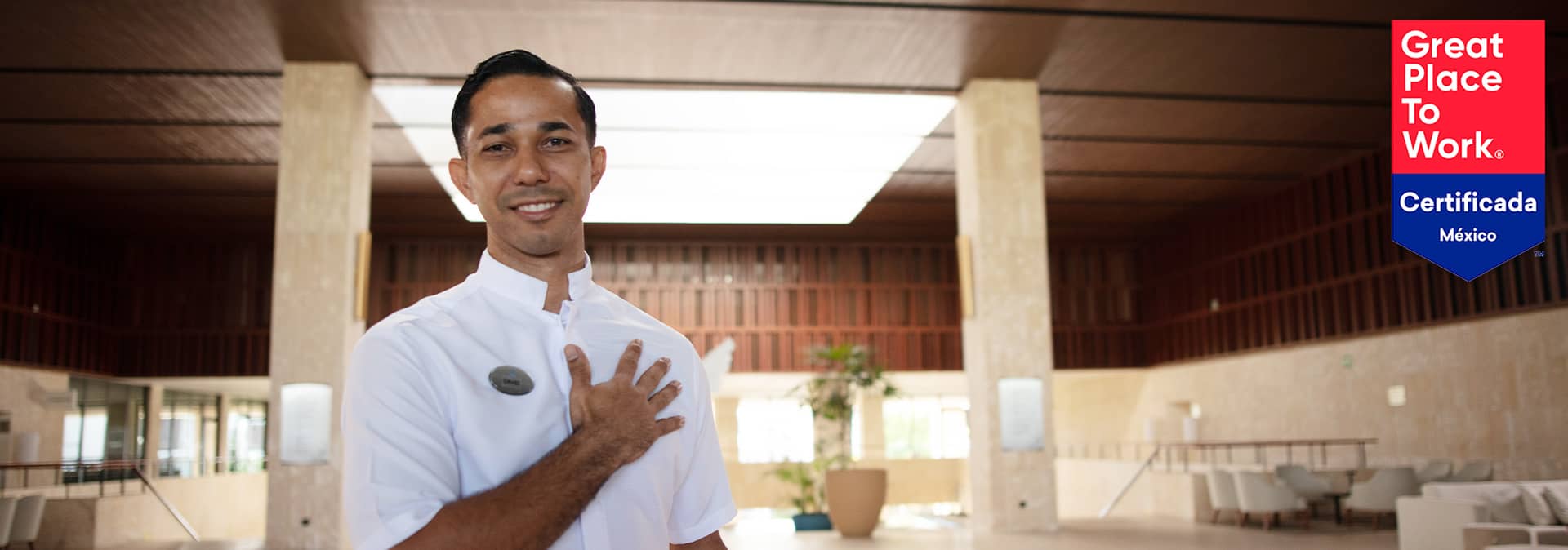 A smiling hotel staff member in a white uniform stands with a hand over his heart in a spacious, modern lobby—ideal for meetings and conference events. A Great Place to Work Certificada México badge is displayed in the top right corner.
