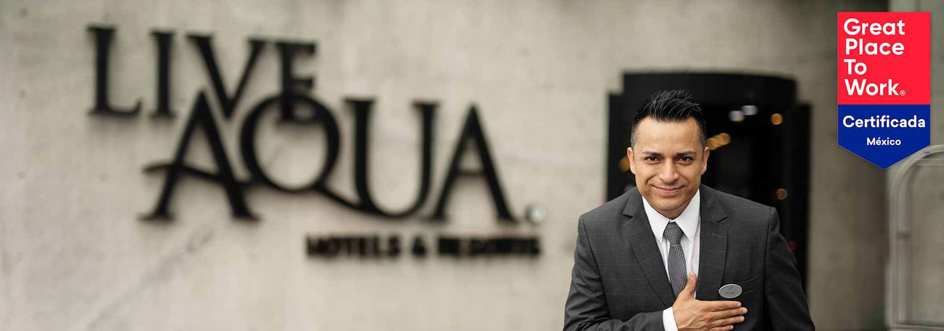 A smiling hotel staff member in a suit stands with his hand on his chest in front of a Live Aqua Hotels & Resorts sign, highlighting the excellence of Mexico hotels for meetings and conference events. A red and blue Great Place to Work Certificada México badge is shown on the right.
