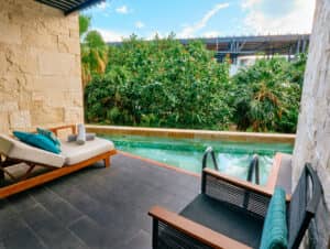 Un patio privado con una tumbona y una silla negra ofrece vistas a una pequeña piscina rodeada de exuberantes plantas tropicales, perfecta para relajarse después de eventos de conferencias en los complejos turísticos de México, enmarcada por muros de piedra bajo un cielo brillante.