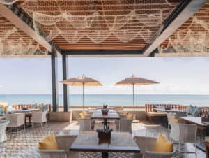 Restaurante al aire libre junto al mar con mesas de madera, sillas acolchadas y una red decorativa en el techo. Ideal para conferencias o reuniones, el comedor de este resort mexicano ofrece vistas a un tranquilo océano azul bajo un cielo despejado.
