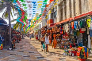 Un animado mercado al aire libre cerca de los centros turísticos de México, con coloridas banderas ondeando y puestos repletos de ropa, sombreros y recuerdos. Una mujer vestida de blanco pasea en bicicleta por la calle empedrada, bordeada de palmeras y compradores.